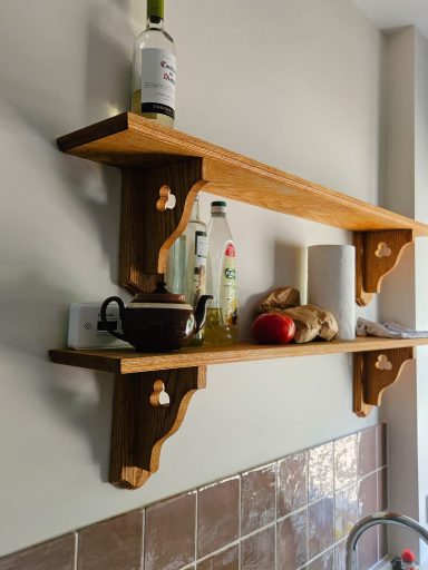 Wooden shelves holding a bottle, oil, a cup, bread, and a red apple against a beige wall.