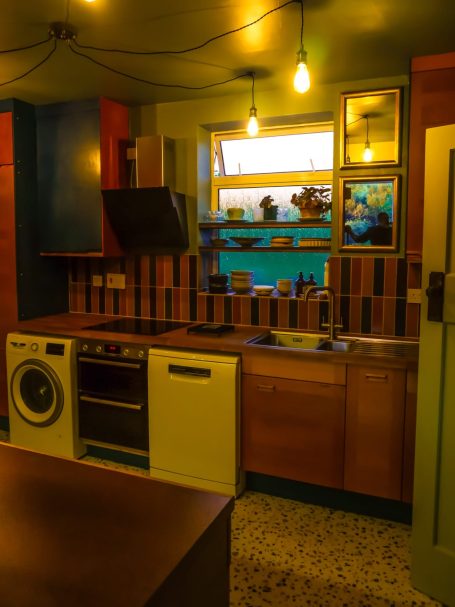 Modern kitchen with wooden cabinets, appliances, and colourful walls, softly lit by hanging bulbs.